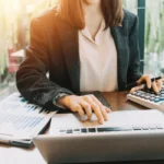 A businesswoman working on financial calculations with a laptop and calculator in a professional setting.