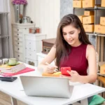 A young woman running an online business from home, packaging products while using her smartphone and laptop.